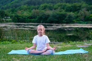 mooi gelukkig meisje met Gesloten ogen beoefenen yoga in de lotus positie in de buurt de meer in natuur. foto