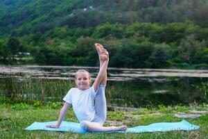 de weinig meisje verhoogt haar been omhoog, gymnastiek opdrachten in natuur. flexibel kind, fitheid, uitrekken, yoga, sport, aan het leren, actief levensstijl concepten foto