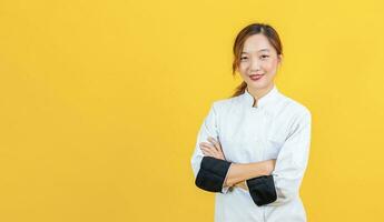 portret studio schot van Aziatisch vrouw chef met professioneel uniform poseren met kruis hand- in zelfverzekerd geïsoleerd Aan geel achtergrond voor culinaire en restaurant concept. foto