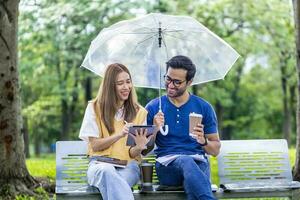 jonge Aziatische en Indiase koppels genieten van ontspannende tijd in de regen samen in het openbare park terwijl ze samen op de bank zitten tijdens het weekend foto