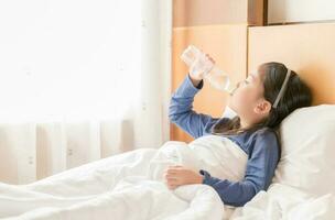 schattig weinig meisje drinken water Aan bed in ochtend, gezond concept foto