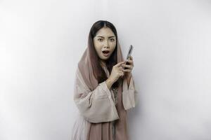 geschokt Aziatisch vrouw vervelend hoofddoek, Holding haar telefoon, geïsoleerd door wit achtergrond foto