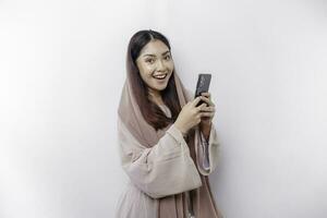 een gelukkig Aziatisch moslim vrouw vervelend een hoofddoek, Holding haar telefoon, geïsoleerd door wit achtergrond foto