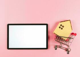 vlak lay-out van digitaal tablet en houten huis model- in boodschappen doen trolley Aan roze achtergrond met kopiëren ruimte, huis aankoop concept. foto