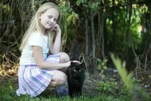 weinig blond meisje Toneelstukken met een zwart kat Aan een achtergrond van groen struiken. foto