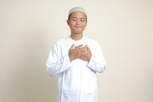 portret van aantrekkelijk Aziatisch moslim Mens in wit overhemd met kalotje plaatsen hand- Aan hart, gevoel heel dankbaar. geïsoleerd beeld Aan grijs achtergrond foto