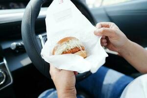 Aziatisch vrouw bestuurder houden en eten Hamburger in auto, gevaarlijk en risico een ongeluk. foto