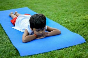 Aziatisch slim kind aan het doen yoga houding in de maatschappij park buitenshuis, kinderen yoga houding. de weinig jongen aan het doen yoga en meditatie oefening. foto