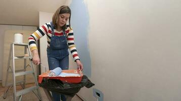 vrouw gekleed in overall en gestreept blouse, gezien van de voorkant, dompelen een rol in de dienblad met blauw verf, binnen een leeg kamer met wit muren voor de helft geschilderd met blauw verf foto