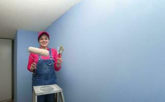 vrouw gekleed in overall en rood t-shirt staand Aan een metaal ladder, met borstels en een rol in haar handen, op zoek Bij de camera, De volgende naar een blauw muur in een leeg kamer foto