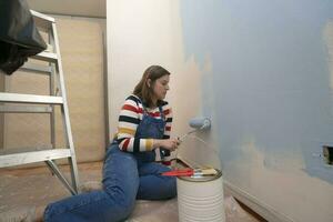 vrouw gekleed in overall en gestreept blouse, profiel visie, schilderij een wit muur met een rol met blauw verf, binnen een leeg kamer met een metaal ladder, verf pot en borstels foto