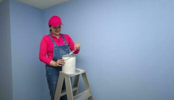 vrouw gekleed in overall en rood t-shirt staand Aan een metaal ladder dompelen haar borstel in een pot van blauw verf De volgende naar een blauw muur foto