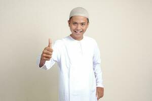 portret van aantrekkelijk Aziatisch moslim Mens in wit overhemd tonen duim omhoog, mooi zo baan hand- gebaar. geïsoleerd beeld Aan grijs achtergrond foto