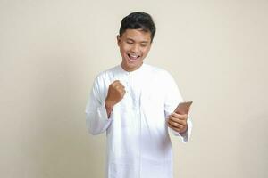 portret van aantrekkelijk Aziatisch moslim Mens in wit overhemd verhogen zijn vuist, vieren winnend spel of krijgen mooi zo nieuws Aan zijn mobiel telefoon. geïsoleerd beeld Aan grijs achtergrond foto