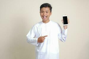 portret van aantrekkelijk Aziatisch moslim Mens in wit overhemd tonen en presenteren blanco scherm mobiel telefoon. reclame concept. geïsoleerd beeld Aan grijs foto