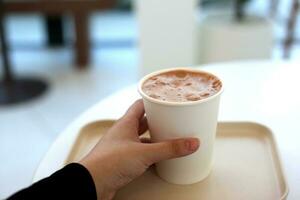 cacao melk papier kop Holding in vrouw hand- met wit tafel in cafe foto