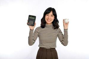 portret van jong Aziatisch vrouw gewoontjes uniform Holding wit varkentje bank en rekenmachine geïsoleerd Aan wit achtergrond, financieel en bank besparing geld concept foto