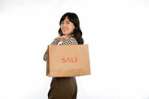 gelukkig jong Aziatisch vrouw gewoontjes uniform Holding boodschappen doen papier zak geïsoleerd Aan wit achtergrond. boodschappen doen korting concept foto