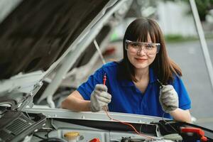 vrouw auto monteur opent auto kap naar inspecteren motor schade en uitvoeren professioneel onderhoud hij droeg een blauw uniform en geïnspecteerd en gerepareerd zijn auto in de werkplaats. foto