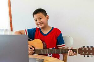 de verhaal van een jongen aan het kijken een notitieboekje computer terwijl voorbereidingen treffen naar praktijk spelen gitaar Bij huis. jongens nemen klassiek gitaar lessen online. foto