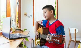 de verhaal van een jongen aan het kijken een notitieboekje computer terwijl voorbereidingen treffen naar praktijk spelen gitaar Bij huis. jongens nemen klassiek gitaar lessen online. foto