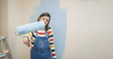 rol met blauw verf in de hand- van een staand vrouw gekleed in overall en gestreept blouse, gezien van de voorkant, tegen achtergrond van half geschilderd wit muur met beroertes van blauw verf foto