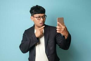 portret van jong Aziatisch bedrijf Mens in gewoontjes pak met verrast uitdrukkingen richten vinger Bij de telefoon. geïsoleerd beeld Aan blauw achtergrond foto