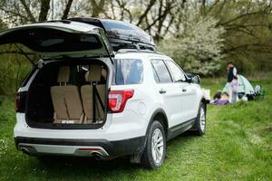 groot suv auto met Open romp en dak rek doos tegen gelukkig jong familie hebben pret en genieten van buitenshuis Aan picknick deken Bij tuin voorjaar park. foto