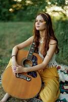 hippie eco vriendelijk vrouw met een gitaar ontspannende in natuur zittend Aan een plaid door de meer in de avond in de stralen van de instelling zon. een levensstijl in harmonie met de lichaam en natuur foto
