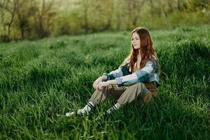 een jong mooi vrouw zit Aan de groen gras in de park en looks uit in de instelling zon foto