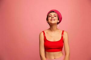 jong atletisch vrouw met een kort kapsel en Purper haar- in een rood top en een roze hoed met een atletisch figuur glimlacht en grimassen op zoek Bij de camera Aan een roze achtergrond foto
