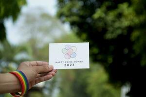 'Vrolijk trots maand 2023' kaart Holding in hand- welke heeft regenboog polsbandje in de omgeving van het, concept voor uitnodigend allemaal mensen naar toetreden de lgbtq evenementen in de omgeving van de wereld in trots maand. foto