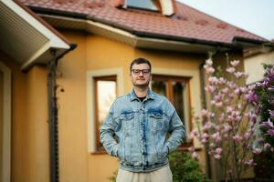 portret van elegant Mens in bril en jeans jasje gesteld tegen zijn huis. foto