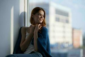 vrouw zittend in de buurt de venster met een blauw plaid dromerig kijken foto