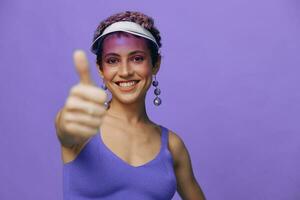 portret van een sportief mode vrouw poseren glimlachen met tanden en richten een vinger Bij de camera in een Purper yoga trainingspak en een transparant pet Aan een Purper monochroom achtergrond foto