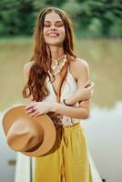 een jong vrouw in een hippie kijken en eco-jurk dansen buitenshuis door de meer vervelend een hoed en geel broek in de zomer zonsondergang foto