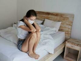 vrouw in een medisch masker in shorts en een t-shirt zit Aan de bed Aan vakantie coronavirus foto