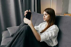 roodharig vrouw met een telefoon in haar hand- zit Aan een sofa en een plaid Aan haar knieën foto