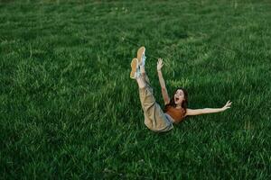 een jong vrouw spelen spellen in de park Aan de groen gras verspreiden haar armen en poten in verschillend routebeschrijving vallend en glimlachen in de zomer zonlicht foto