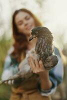 een gelukkig jong vrouw glimlacht net zo ze looks in de camera en houdt een jong kip dat legt eieren voor haar boerderij in de zonlicht. de concept van zorgzaam en gezond gevogelte foto