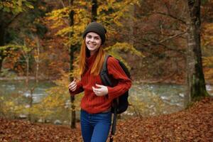 vrouw in herfst park met gedaald bladeren en rugzak Aan haar terug rivier- in de achtergrond foto