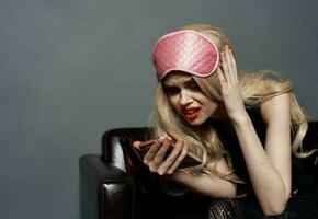 dronken blond vrouw met mobiel telefoon bedenken en roze slaap masker foto