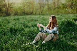 een jong mooi vrouw zit Aan de groen gras in de park en looks uit in de instelling zon foto