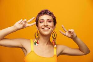 portret van een jong vrouw met een kort kapsel en gekleurde haar- glimlachen en tonen haar tong Bij de camera Aan een oranje achtergrond met oorbellen accessoires in de studio foto