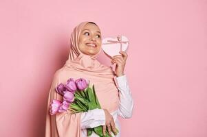 moslim vrouw in roze hijaab, Holding een boeket van Purper tulpen en een hart vormig geschenk doos, glimlachen over- roze achtergrond foto