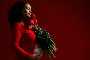 verrukkelijk zwanger vrouw met Gesloten ogen, strelen haar buik, Holding een boeket van rood tulpen. gelukkig moeder dag. foto