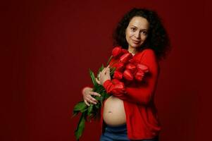 multi etnisch mooi zwanger vrouw met een schattig boeket van tulpen, poseren kaal buik Aan geïsoleerd rood kleur achtergrond. foto
