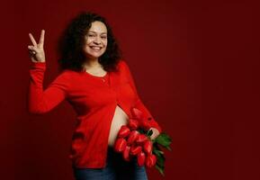 mooi zwanger vrouw met bundel van tulpen, glimlacht, gebaren met vrede teken over- rood achtergrond. gelukkig moeder dag foto