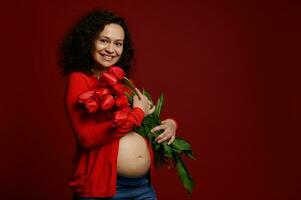 mooi Afrikaanse Amerikaans gelukkig zwanger vrouw glimlachen breed, poseren kaal buik met rood tulpen Aan kleur achtergrond. foto