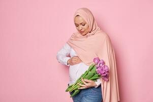 moslim zwanger vrouw in roze hijaab, accenten haar buik, voelt eerste baby trappen, houdt mooi tulpen, geïsoleerd Aan roze foto